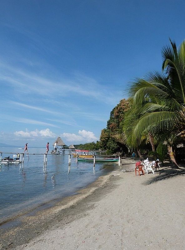 Izabal tropical beach with light gray sand, crystal clear turquoise waters, green palm trees on rocky cliffs and colorful fishing boats