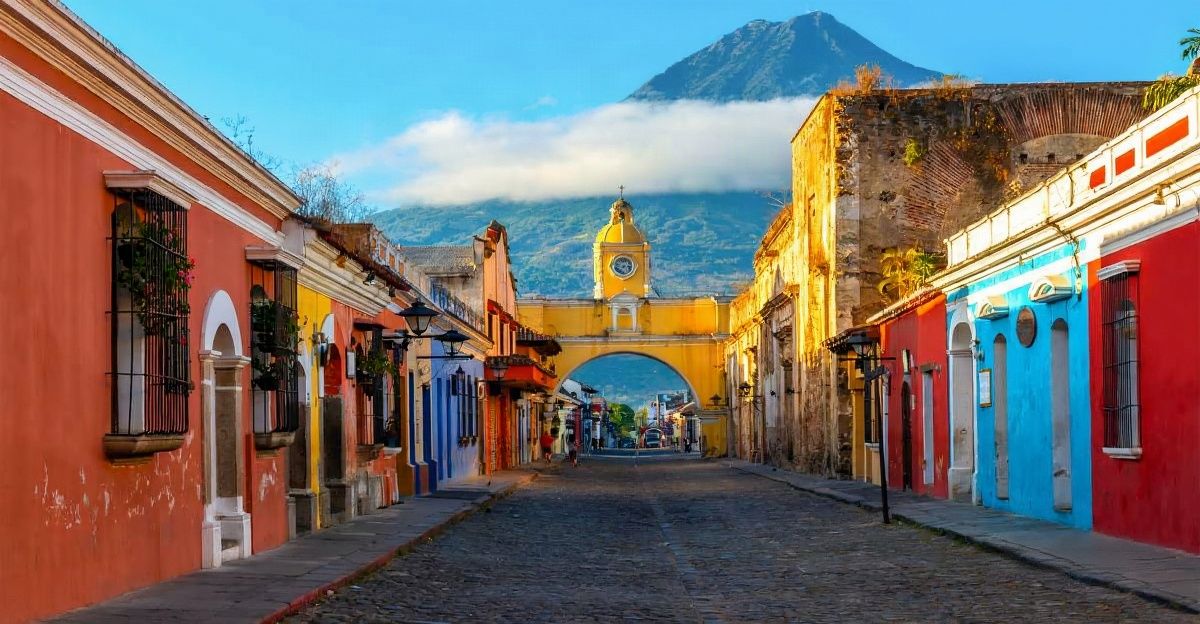Colonial cobblestone street in Antigua Guatemala with colorful baroque buildings, whitewashed stone arches, Agua volcano in the distance and yellow ar