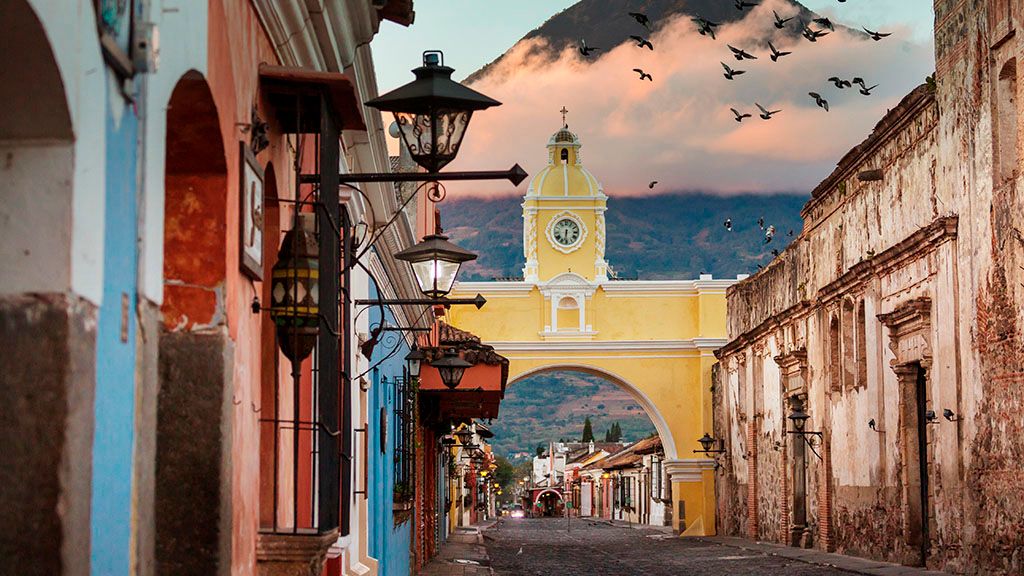 Destinos turísticos como el lago de Atitlán, Xela, la costa del Pacífico y Semuc Champey, Antigua es una primera parada popular cuando se viaja por el país y la región.

En Antigua, puede encontrarlo todo: caminatas al volcán, clases de cocina, recorridos de café, oportunidades de voluntariado, vida nocturna local y mucho más. Estas son algunas de las principales atracciones y recorridos por descubrir de Antigua:

## Parque Central

En el corazón de Antigua, se encuentra el parque central o Central Park Plaza. Un hervidero de actividad y encanto colonial, la Plaza Central Park es el centro de la ciudad y la vida de los antigueños. El parque es el lugar perfecto para pasear tranquilamente, observar a los lugareños mientras pasan sus días o disfrutar de un café en los cafés vecinos.

## Arco de Santa Catalina

El Arco de Santa Catalina es el arco amarillo icónico de Antigua. Construido en 1694, el arco servía de puente entre conventos permitiendo a las monjas cruzar la calle sin ser vistas. Hoy en día, el arco proporciona un colorido telón de fondo para las fotos (asegúrese de obtener uno con el Volcán de Agua de fondo) y es una atracción básica en los recorridos a pie de Antigua.

## Iglesia La Merced

Antigua tiene muchas iglesias impresionantes, pero la Iglesia La Merced podría ser la más bonita de todas. Esta iglesia de estilo barroco bien conservada tiene una impresionante fachada amarilla con estuco blanco ornamentado, lo que la convierte en uno de los sitios más reconocibles de Antigua. También puede visitar las ruinas del convento de al lado por una pequeña tarifa de entrada.

## Cerro de la Cruz

Situado al norte de Antigua se encuentra el Cerro de la Cruz (Cerro de la Cruz). A una corta caminata de media hora o de fácil acceso en automóvil o tuk-tuk, Cerro de la Cruz ofrece vistas panorámicas de Antigua y sus pueblos, colinas y volcanes circundantes. Este lugar es uno de los favoritos tanto de los lugareños como de los visitantes. La vista se disfruta mejor por la mañana para obtener cielos despejados y vistas ininterrumpidas del espectacular paisaje.

## Mercado local de Antigua

Al caminar por Antigua, encontrará mercados artesanales en casi todos los rincones. El mercado local, sin embargo, es verdaderamente auténtico. Aquí, dentro de un laberinto de puestos, encontrará vendedores que venden productos, artículos para el hogar, ropa de segunda mano, trabajos en madera y mucho más. Intente visitar en un día de mercado (lunes, jueves o sábado) para disfrutar de la experiencia completa.

## Aprende español

Antigua es un lugar famoso para aprender español y los visitantes pueden elegir entre una gran cantidad de escuelas alrededor de la ciudad colonial. La mayoría de las escuelas imparten clases individuales y los precios son accesibles para la mayoría de los presupuestos. Ya sea que desee un curso intensivo de español de supervivencia para sus viajes o esté buscando estudiar un idioma más serio, Antigua es EL destino.

## Aprenda a bailar salsa

Antigua tiene una escena de baile de salsa decente y para aquellos que buscan diversión nocturna e inmersión cultural, muchos de los estudios de baile de Antigua ofrecen clases gratuitas durante la semana para principiantes. Si viaja solo, las clases también son una excelente manera de conocer a otros viajeros y lugareños por igual.

## Caminatas por volcanes

Antigua está rodeada de volcanes y se puede acceder fácilmente desde Antigua a dos de las caminatas por volcanes más famosas de Guatemala: Pacaya y Acatenango. Pacaya ofrece a los visitantes una caminata accesible de medio día con vistas increíbles y un encuentro cercano con un volcán vivo: los visitantes pueden presenciar lava fundida y tostar malvaviscos sobre rocas volcánicas calientes durante el recorrido. Aquellos que buscan una caminata más desafiante, pueden escalar el volcán Acatenango durante la noche y presenciar las magníficas erupciones del cercano Volcán de Fuego y las impresionantes vistas sobre las nubes.